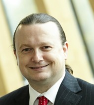 A headshot of Prof. Laurence Ferry, smiling in a suit against a blurred background.